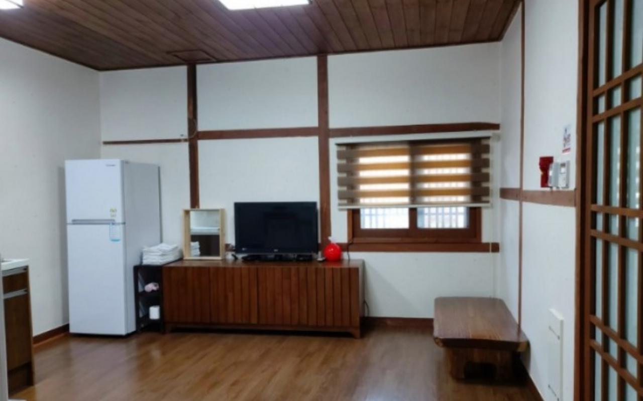 원목 천장과 마룻바닥에 냉장고, TV와 수납장, 좌식 탁자가 놓인 거실 공간입니다.